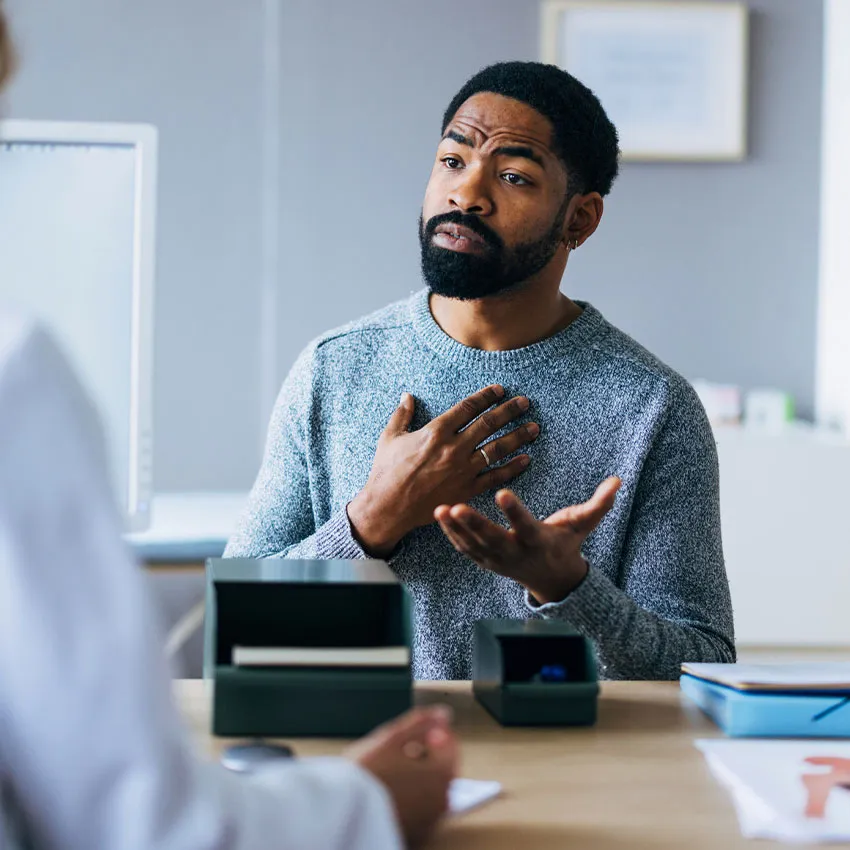 Man in gray sweater speaking earnestly during an office consultation at a desk with documents and a professional