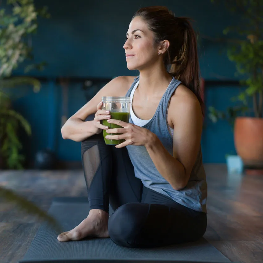 A woman drinking a green smoothie on a yoga mat in a fitness studio, relaxed post-workout wellness moment
