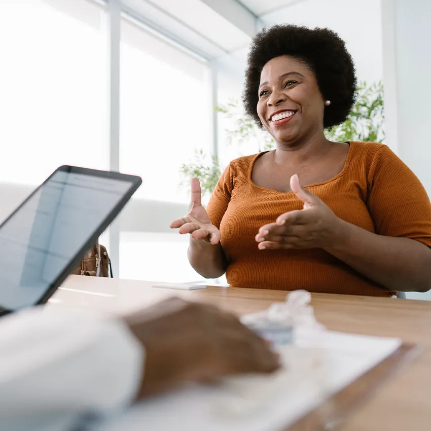 Smiling woman in orange top gesturing during a business meeting in a bright office across a desk with laptop