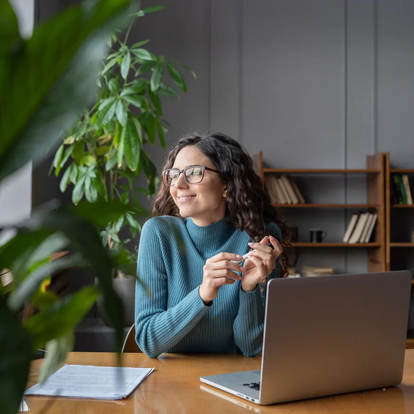 Smiling woman wearing glasses working on laptop in a plant-filled home office, remote work setup with notebook and bookshelves.