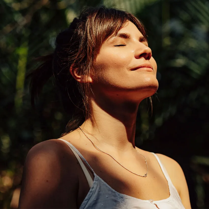 A woman basking in sunlight outdoors with eyes closed, serene expression, white top and necklace