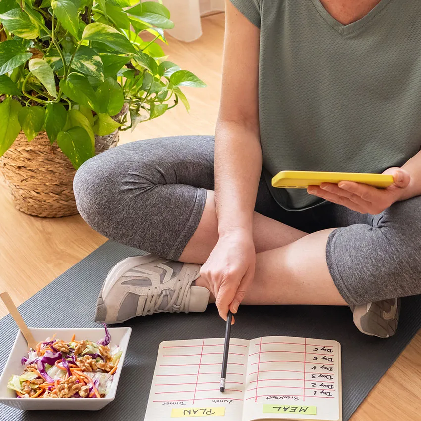 Healthy meal planning: woman on yoga mat writing a meal plan in notebook with smartphone and salad bowl