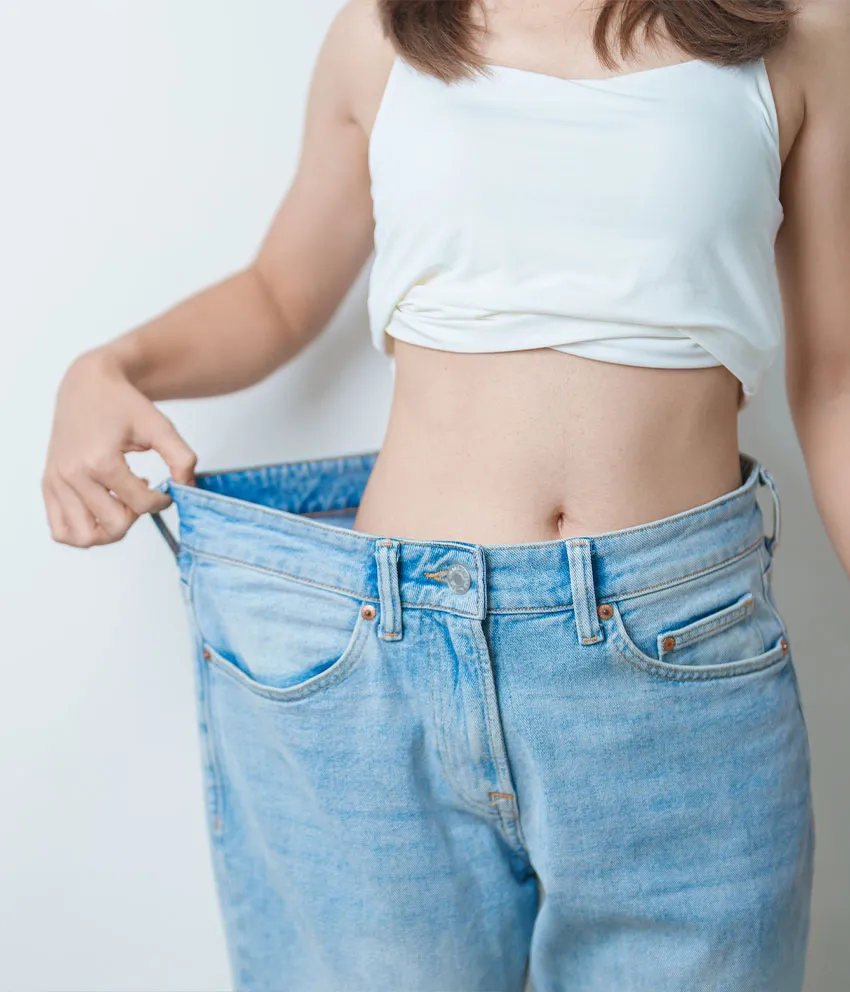 A woman holding out oversized jeans to show weight loss and slimmer waist wearing a white crop top
