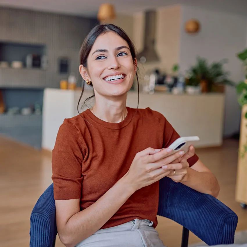 Smiling woman holding smartphone in modern living room, casual brown top, relaxed home lifestyle and digital communication