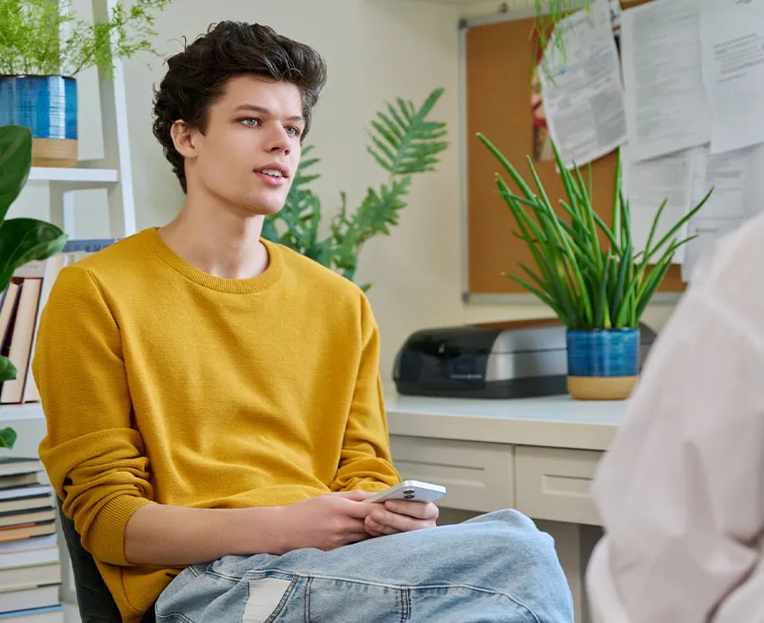 Young man in yellow sweater seated holding smartphone in office with plants, engaged in conversation