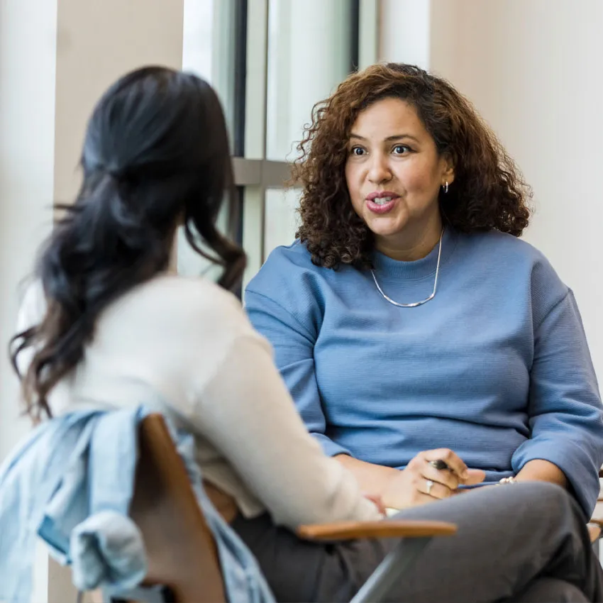 Supportive conversation between two women in an office by a window, one woman listening and taking notes