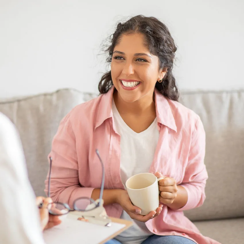 Smiling woman holding a coffee mug on sofa, engaged in a warm conversation in a bright living room