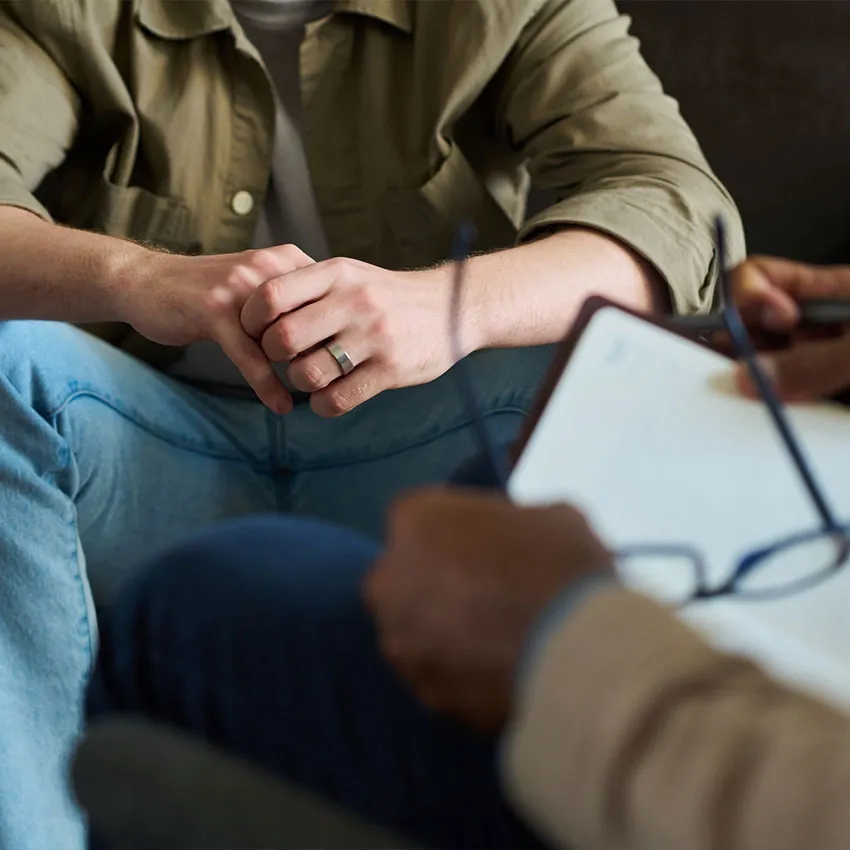 A man with clasped hands in a therapy session while a therapist takes notes on a clipboard