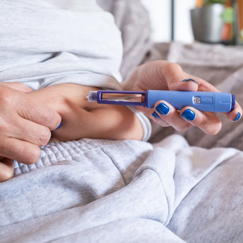 A woman injecting insulin into her abdomen with a blue insulin pen while wearing gray loungewear for diabetes management