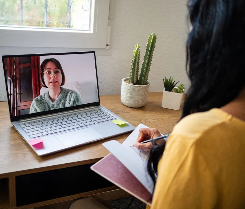 Online video call: woman taking notes on laptop in a home office with potted plants