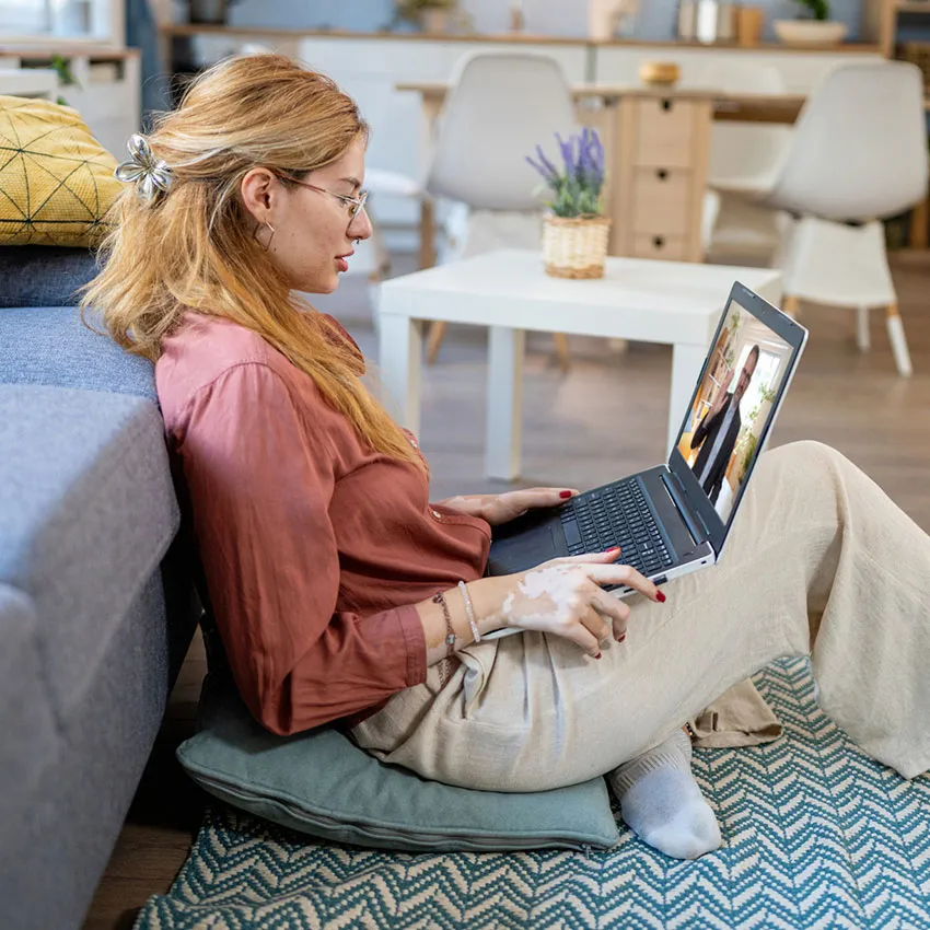 Woman with glasses sitting on a floor cushion using a laptop for a video call in a cozy living room remote work setup.