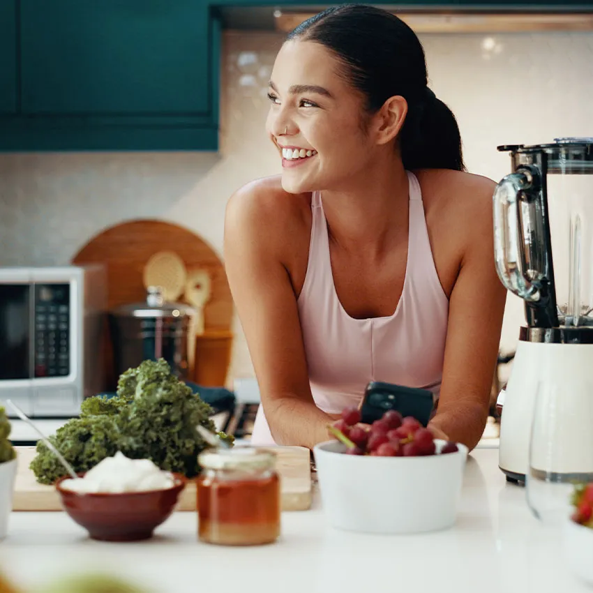 Smiling woman in kitchen with blender, kale, grapes, honey and yogurt preparing a healthy smoothie