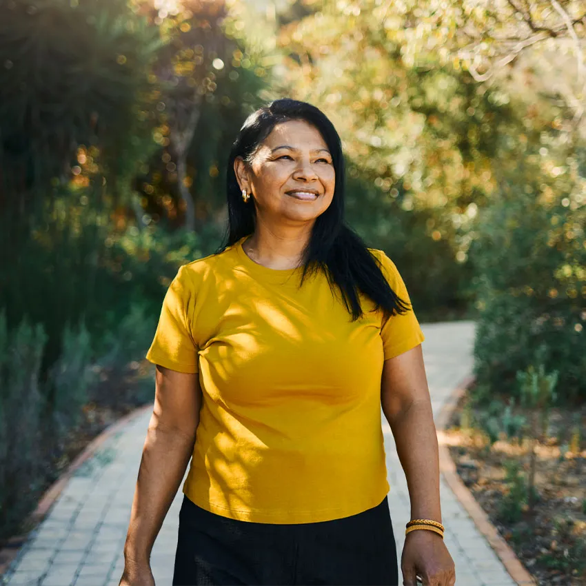 Smiling woman in yellow shirt walking on sunlit park path surrounded by green trees and shrubs