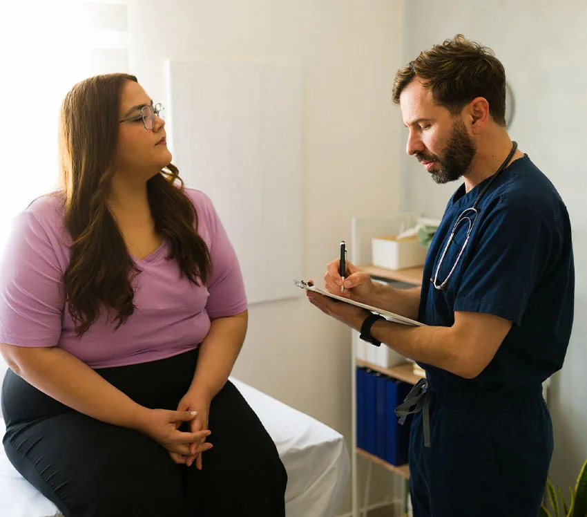 Primary care doctor talking with female patient in exam room, taking notes on clipboard during checkup