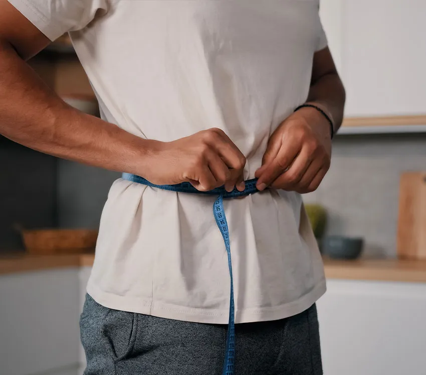 A person measuring waist with blue tape measure at home, tracking body measurements for fitness and weight loss