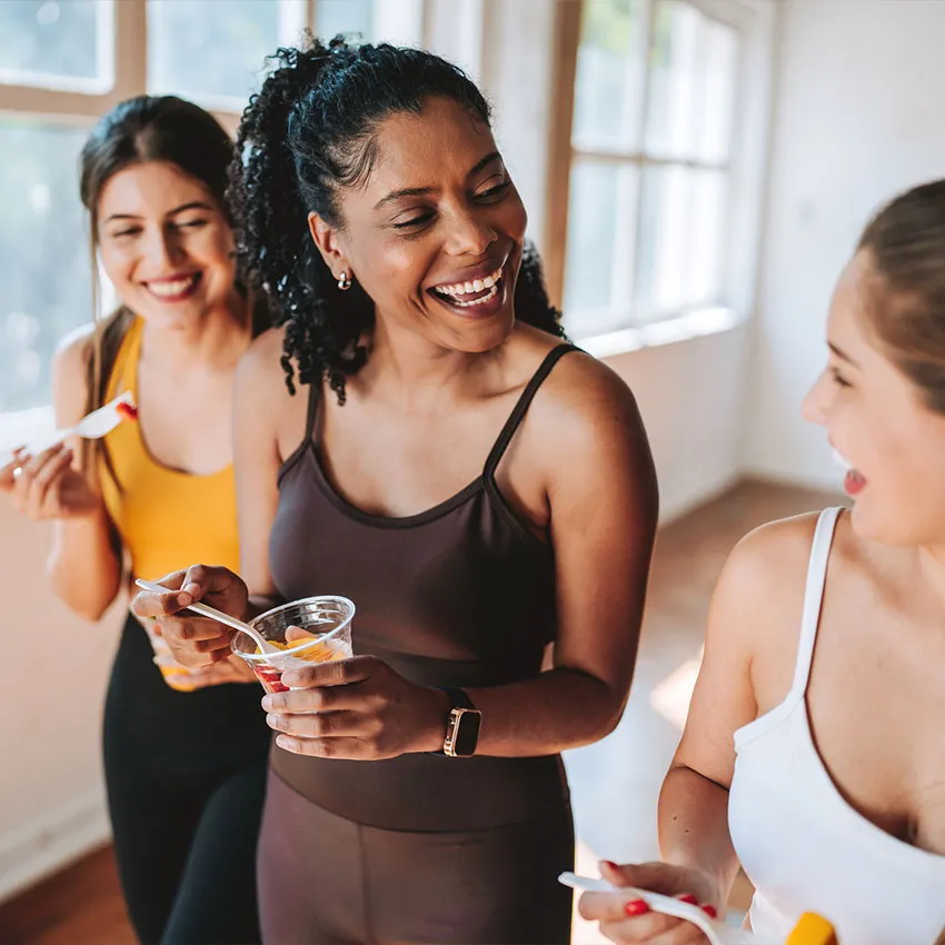 Three women in workout clothes laughing and sharing healthy fruit cups in a sunlit studio, post-workout snack moment
