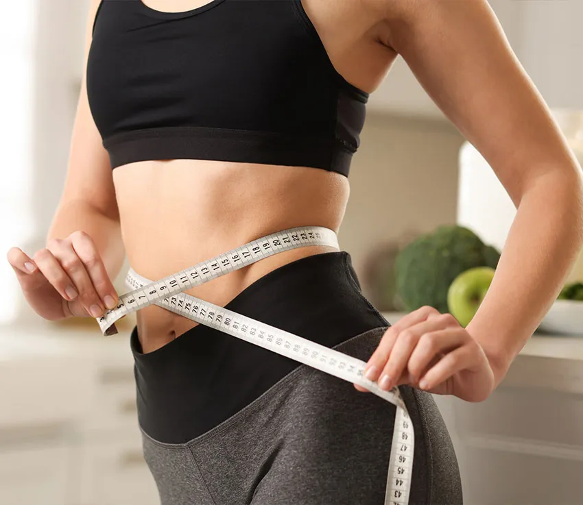 Woman measuring her waist with a tape measure while wearing workout clothes, representing fitness and weight-loss progress.