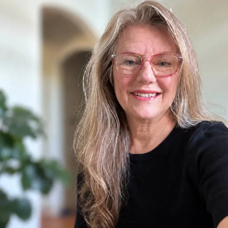 Smiling woman with long gray hair and pink glasses wearing a black top in bright home interior portrait