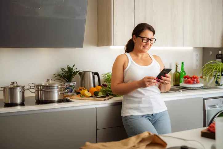 Woman using smartphone in modern kitchen near cutting board with fresh fruits and vegetables, stainless pots on stove.